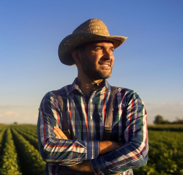 Agricultor com chapéu, de braços cruzados, olhando para o horizonte, com plantações de soja ao fundo.