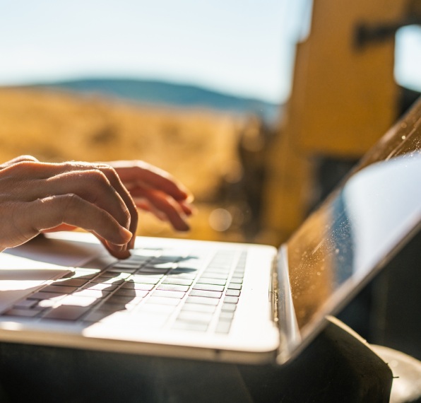 Mãos digitam em um laptop prateado ao ar livre. O fundo, banhado pelo sol, está desfocado